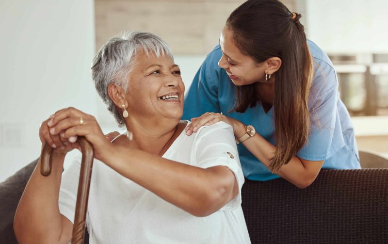 Happy, relax and senior woman with caregiver smile while sitting on a living room sofa in a nursing home. Support, help and professional nurse or healthcare worker helping elderly lady or patient.